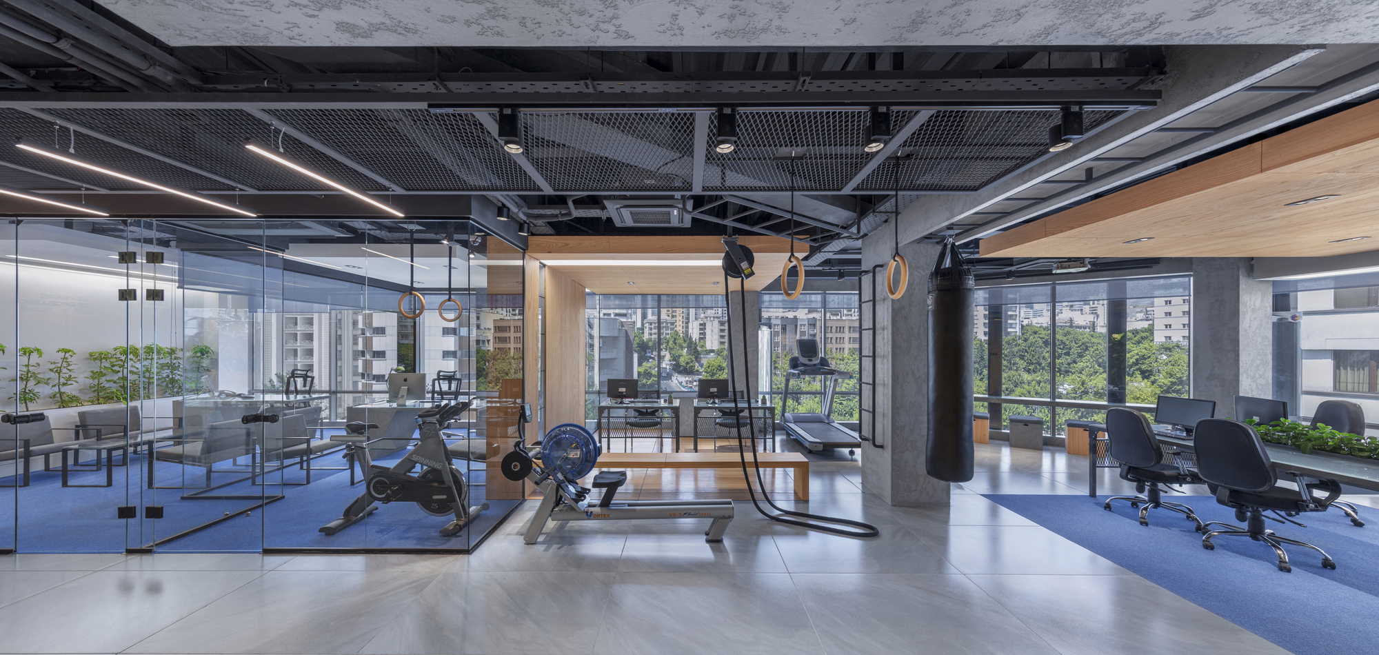 Open office and training floor of AKA Fitness Office, with workout equipment, desks on blue carpet and a black open ceiling.