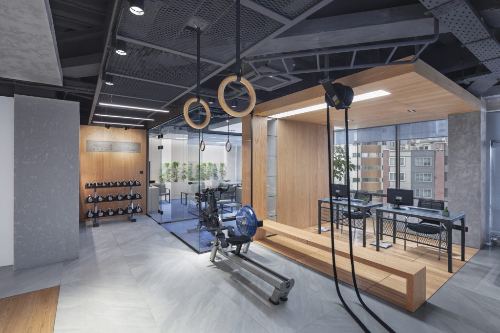 Gym corner with rings, a rowing machine and a wall of dumbbells, next to desks with city views.