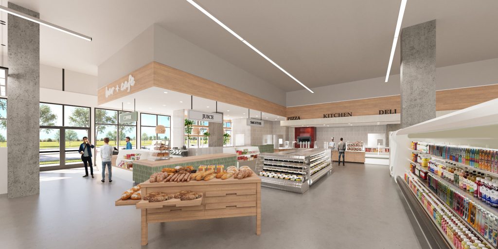 A bakery table of bread in the middle of the store, with deli and kitchen counters under linear lights and stocked refrigerated aisles.