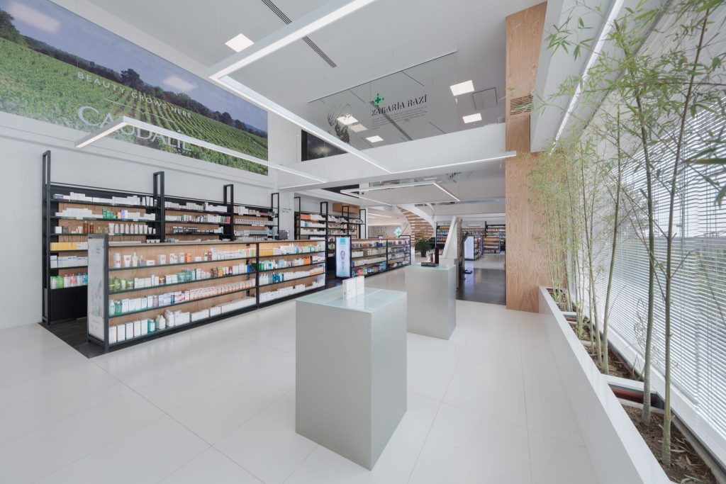 A pharmacy aisle with long white shelving, a double-height space, linear ceiling lights and a row of bamboo planters along the glass.