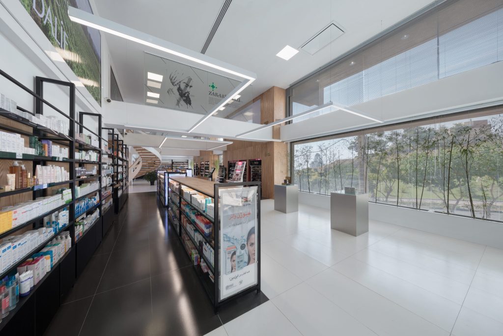 Open pharmacy shelves under a white double-height ceiling, with a glass wall onto trees and a sculptural white stair beyond.