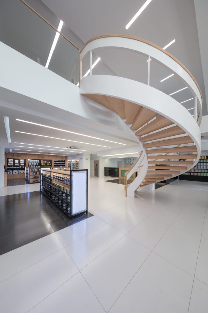 A white spiral stair with a wooden handrail curving up through the double-height pharmacy, beside a glass balustrade.