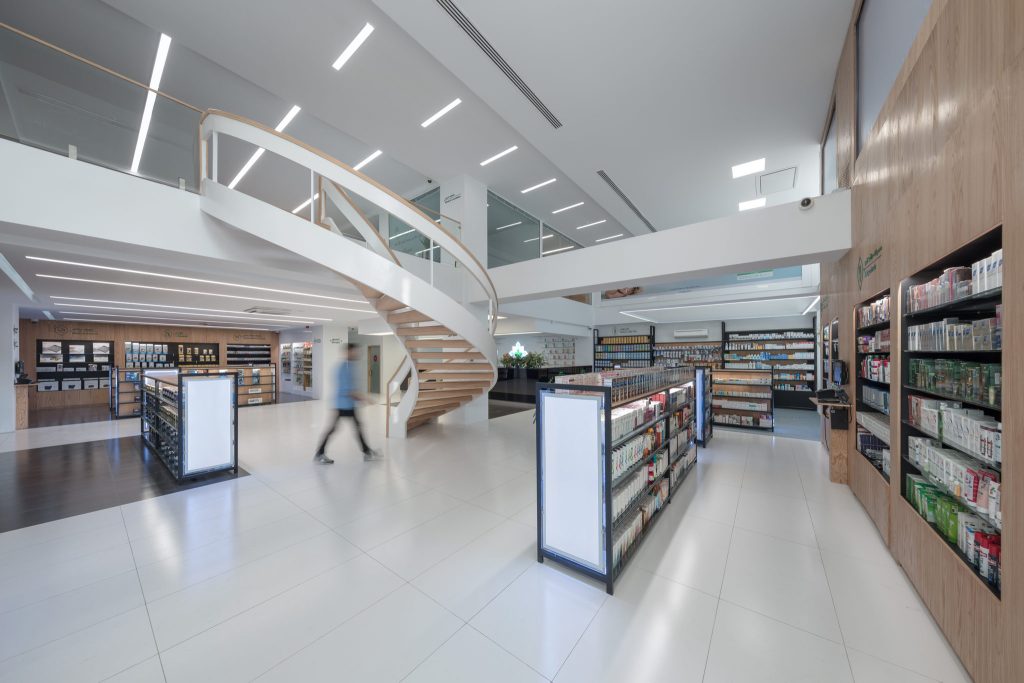 The pharmacy's ground floor with lit display stands, wood-clad walls of shelving and the spiral stair rising to the upper floor.