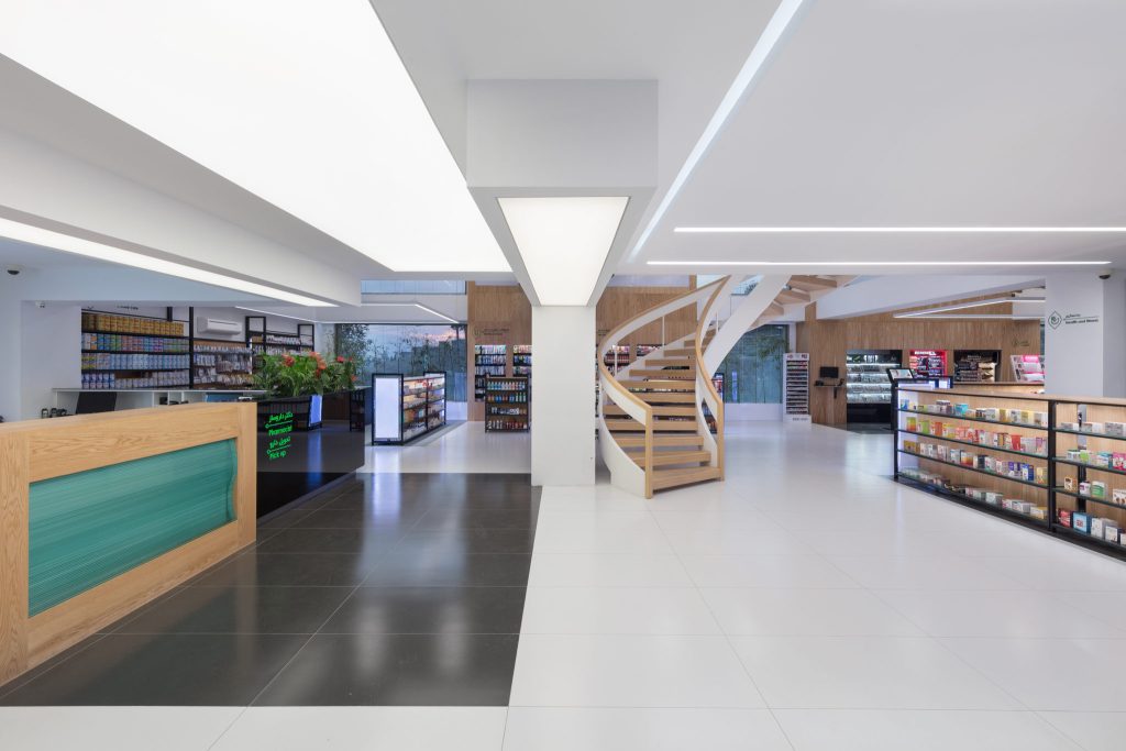 A pharmacy counter with a teal front under a white column, with wood shelving and the spiral stair behind it.