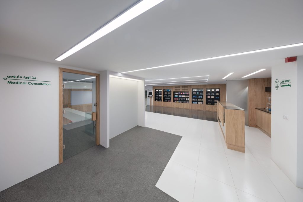 An upstairs corridor with a gray carpet path, a glass-doored consultation room and wood-clad shelving at the end.