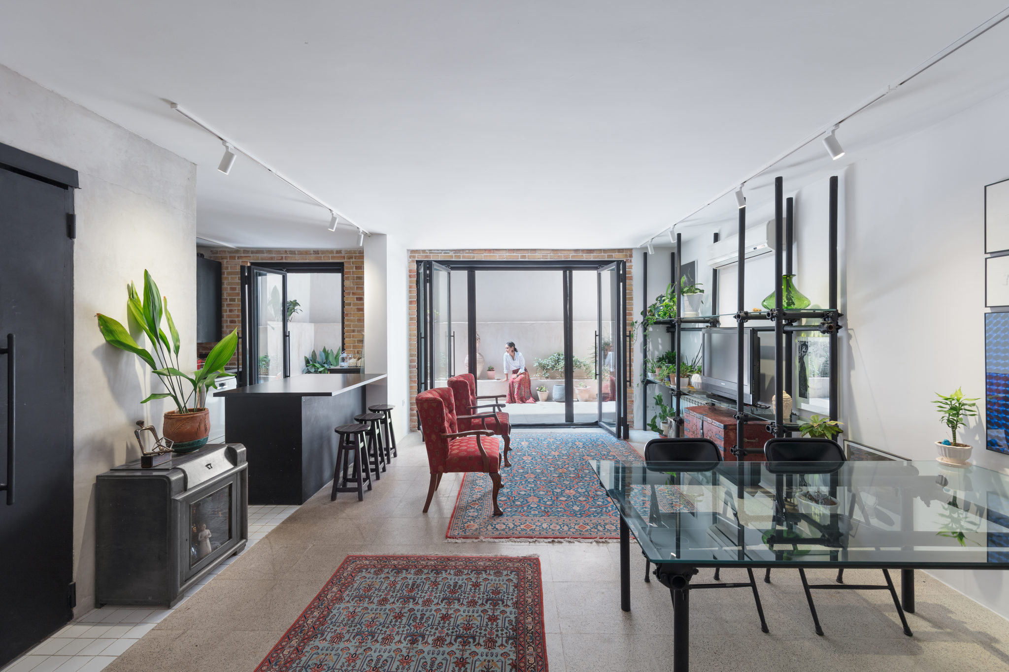 A living room with red armchairs, a Persian rug, a glass dining table and black-framed glass doors onto a raised backyard.