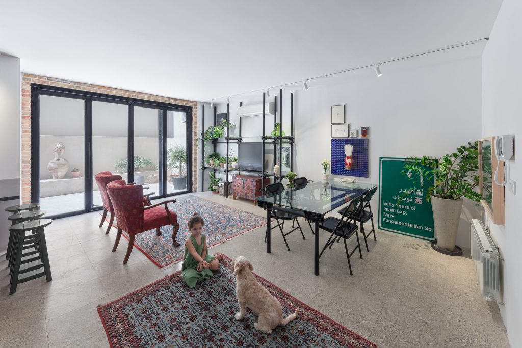 A child and a dog on a Persian rug in a bright living room with red armchairs, plants and a black steel shelf.