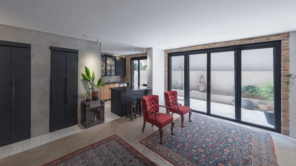 Black metal doors set in a gray plaster wall, red armchairs and a black kitchen island by folding glass doors.
