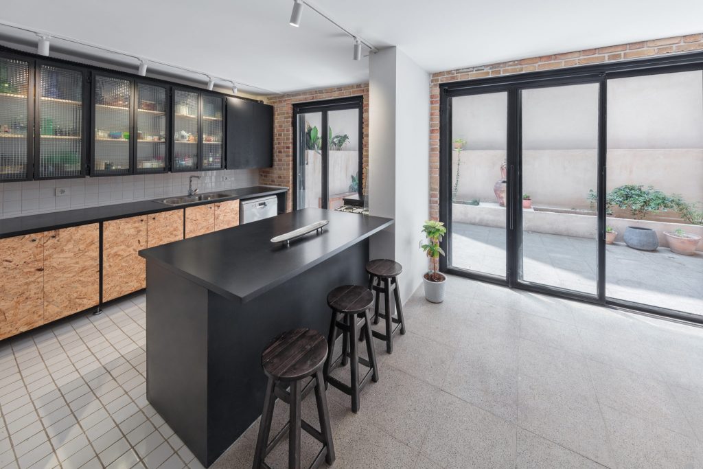 A black kitchen island with wood stools, a white tile floor and a wall of cabinets with wood-panel fronts.