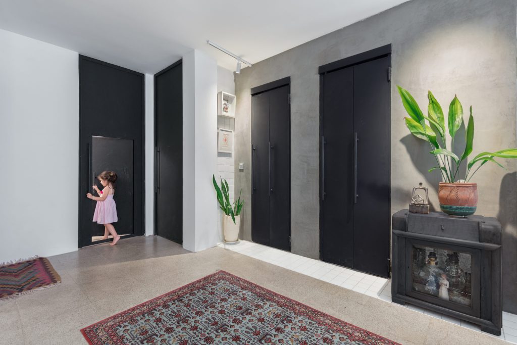 A small child at a child-sized door in a gray plaster wall, beside tall black metal doors and a plant.