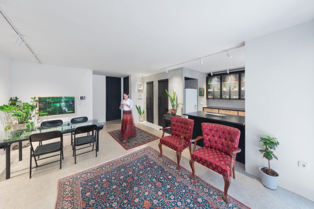 A living and dining room with red armchairs, a Persian rug, a glass table and a woman standing by the kitchen.