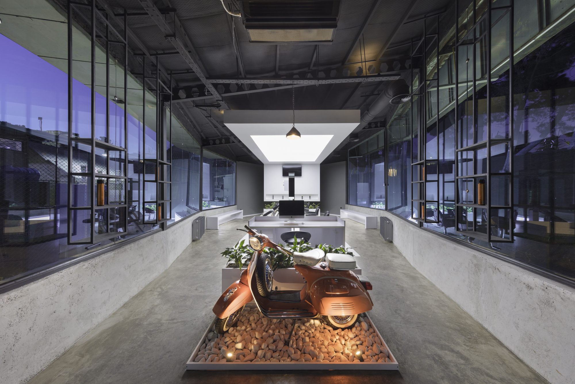 The long, glass-walled hair salon at dusk, with a vintage orange scooter on a pebble display at the entrance and a white lightbox ceiling beyond.