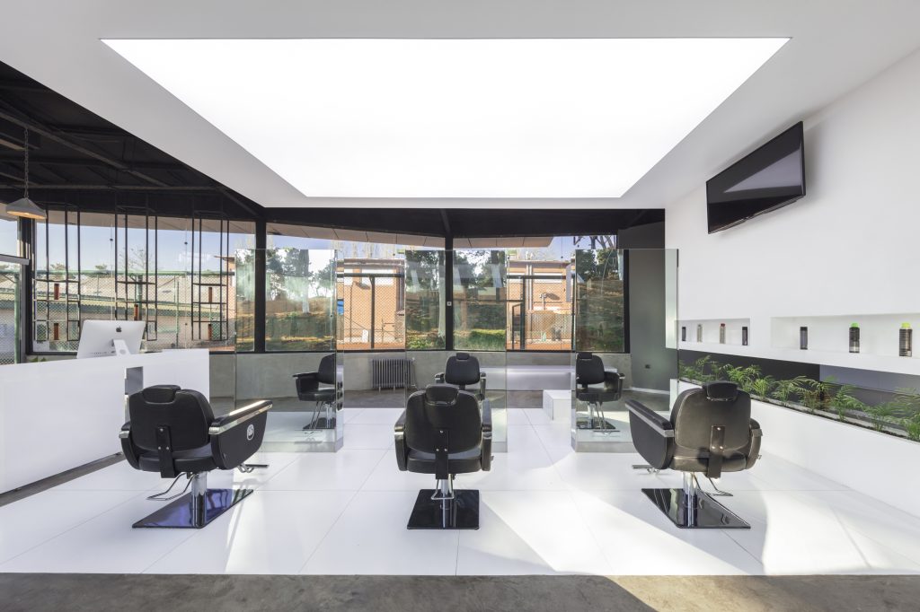Black salon chairs under a glowing white ceiling panel, with glass walls and greenery outside.