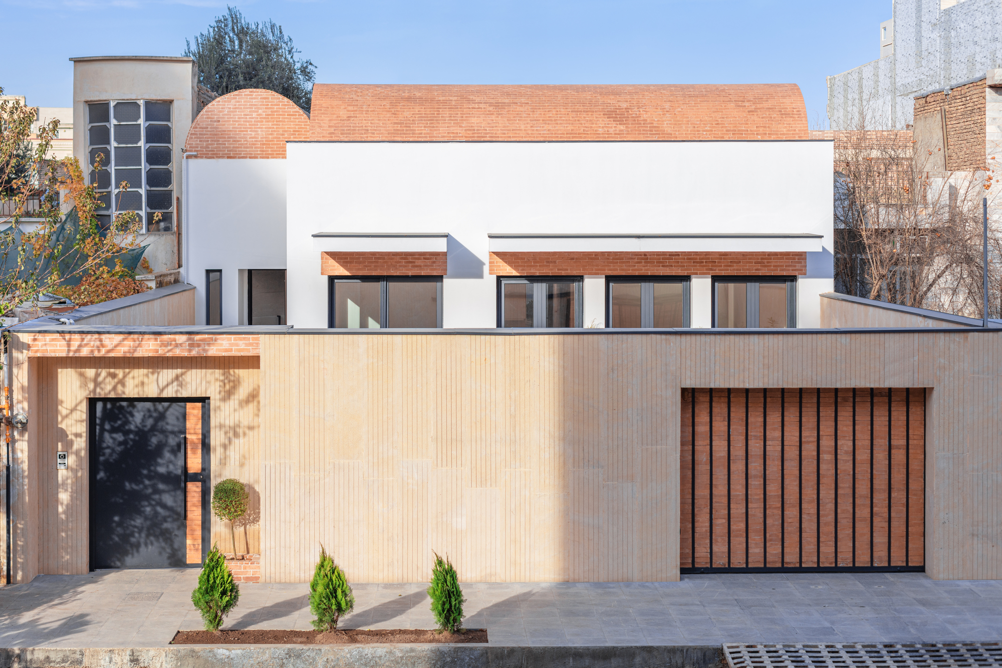 Street front of Vosagh House: a white house with brick barrel-vault roofs behind a beige stone wall, a dark entrance door and a wood-slat garage gate.