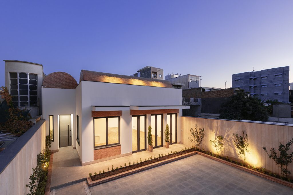 The courtyard of Vosagh House at dusk, with warm light from the living room and a planted edge along the stone wall.
