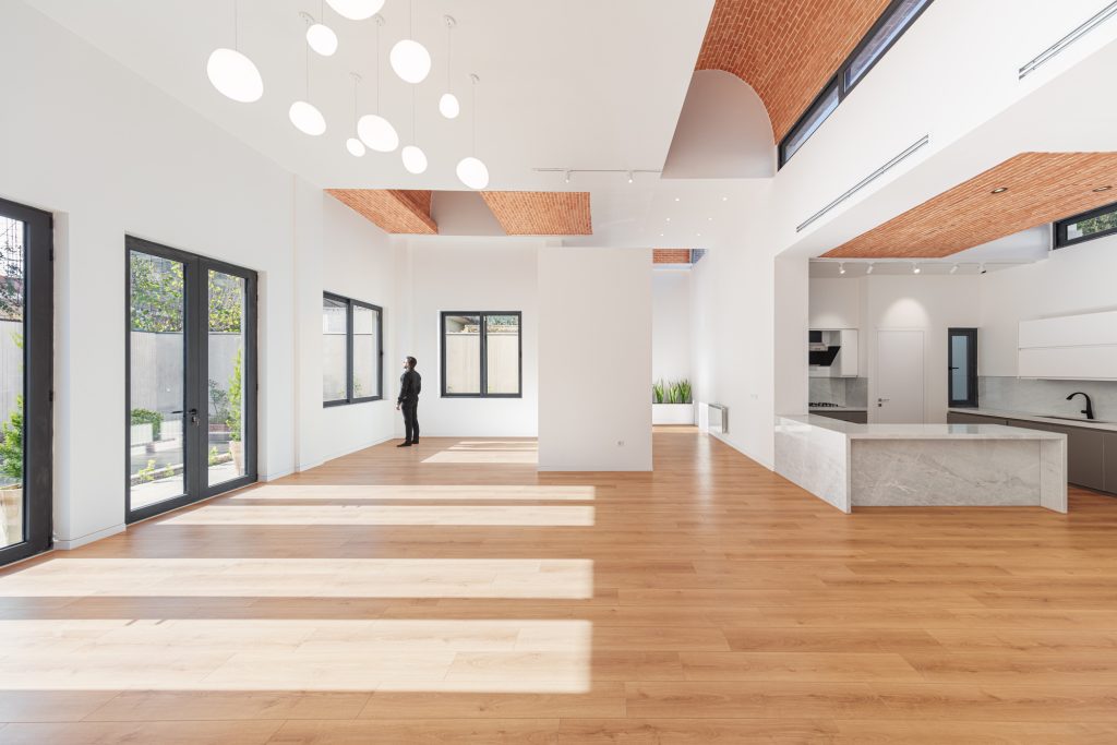 Open living space with a light oak floor, an exposed brick vault, round skylights and the kitchen beyond.